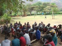 A large group are seated on the ground to receive a health message.